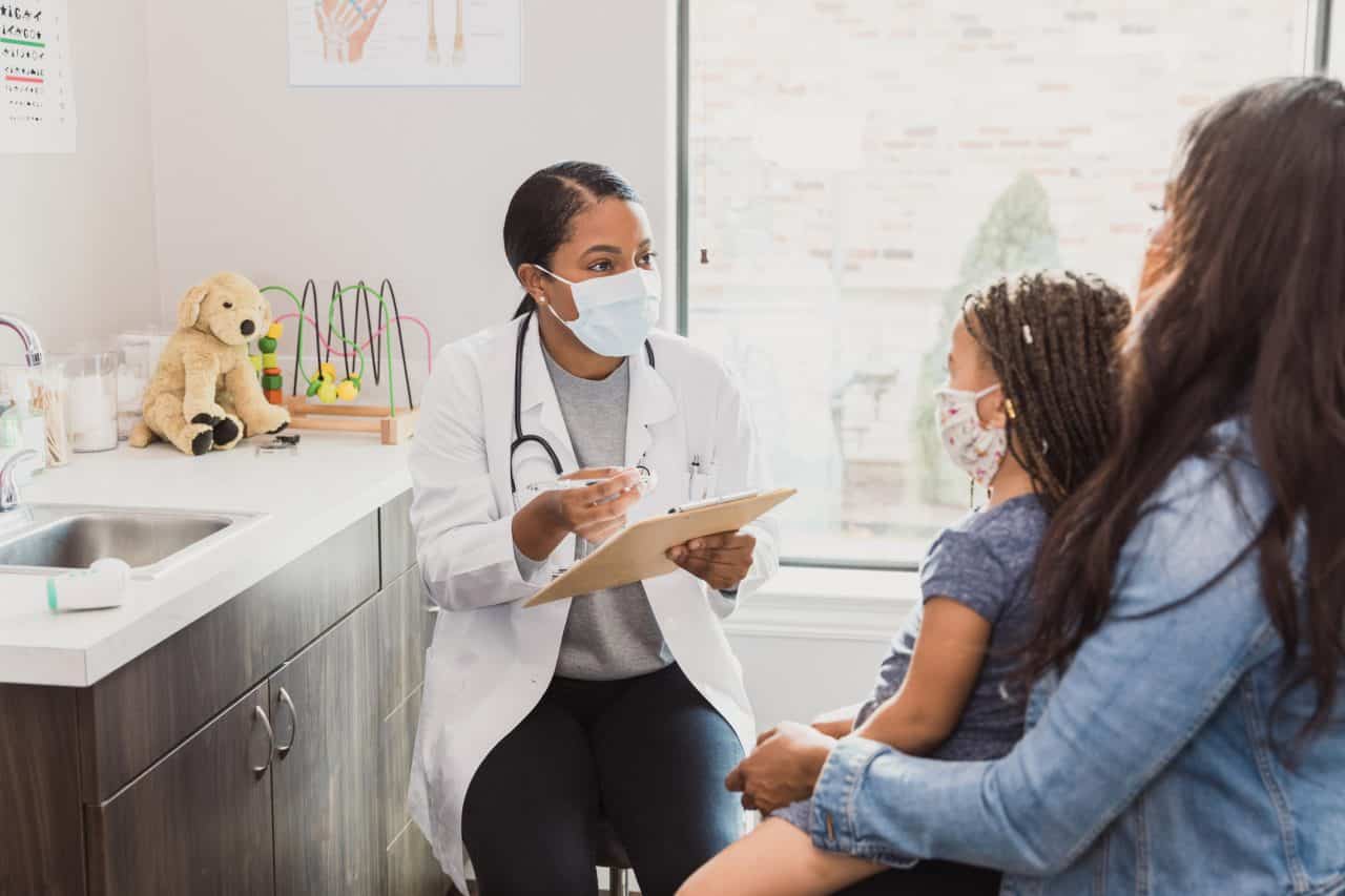 With a protective mask on, a female pediatrician talks to a young patient's mother about the woman's daughter's medical conditions. They are wearing protective masks
