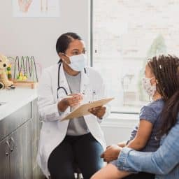 With a protective mask on, a female pediatrician talks to a young patient's mother about the woman's daughter's medical conditions. They are wearing protective masks